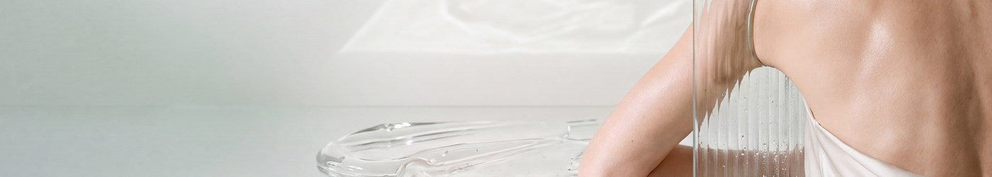 A woman with the frosted OMBRA BODY bottle beside clear glass, in a room of water light.